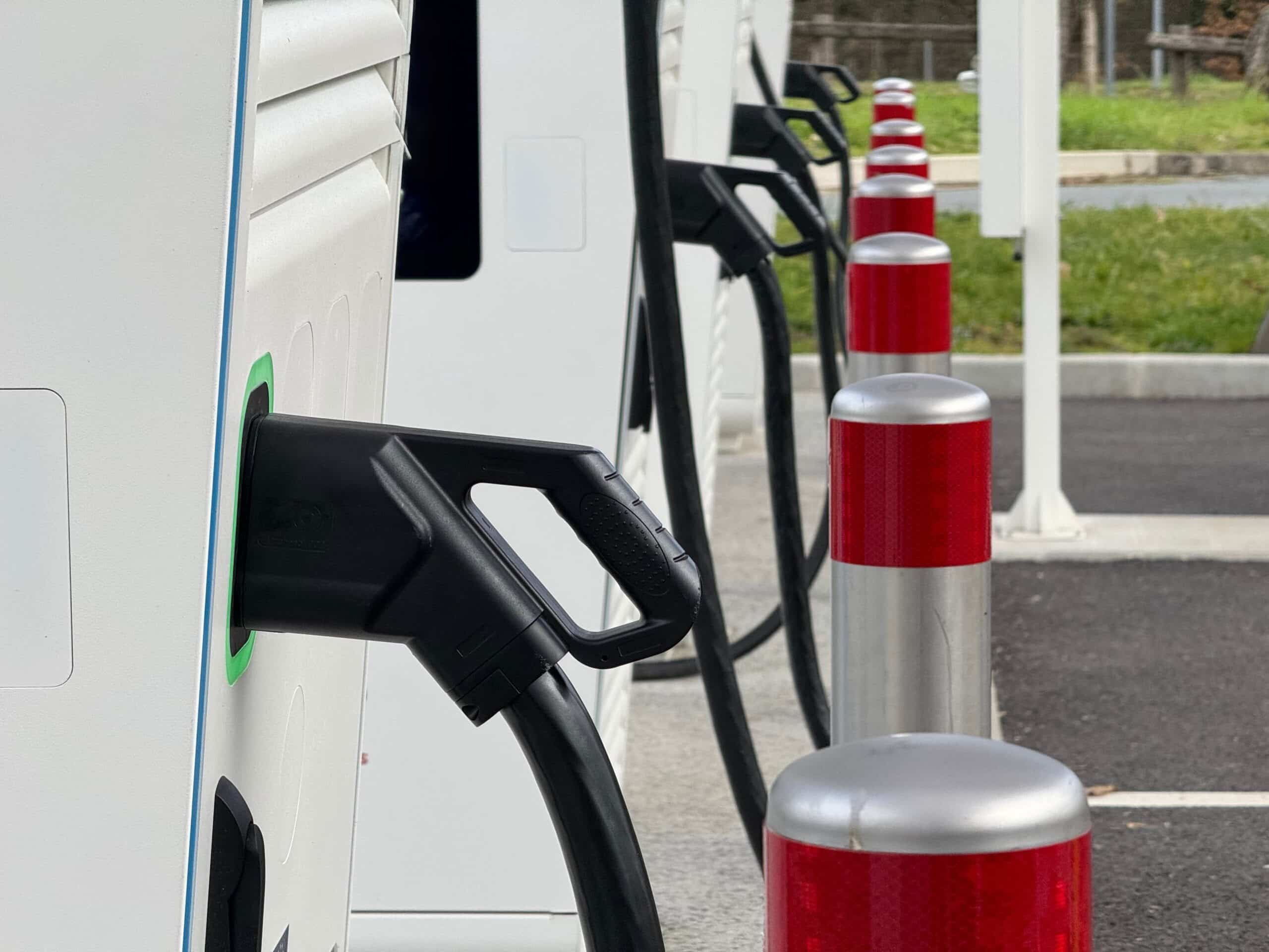 Borne de recharge pour véhicule électrique avec prise branchée. Plusieurs bornes et poteaux rouges alignés dans un parking.