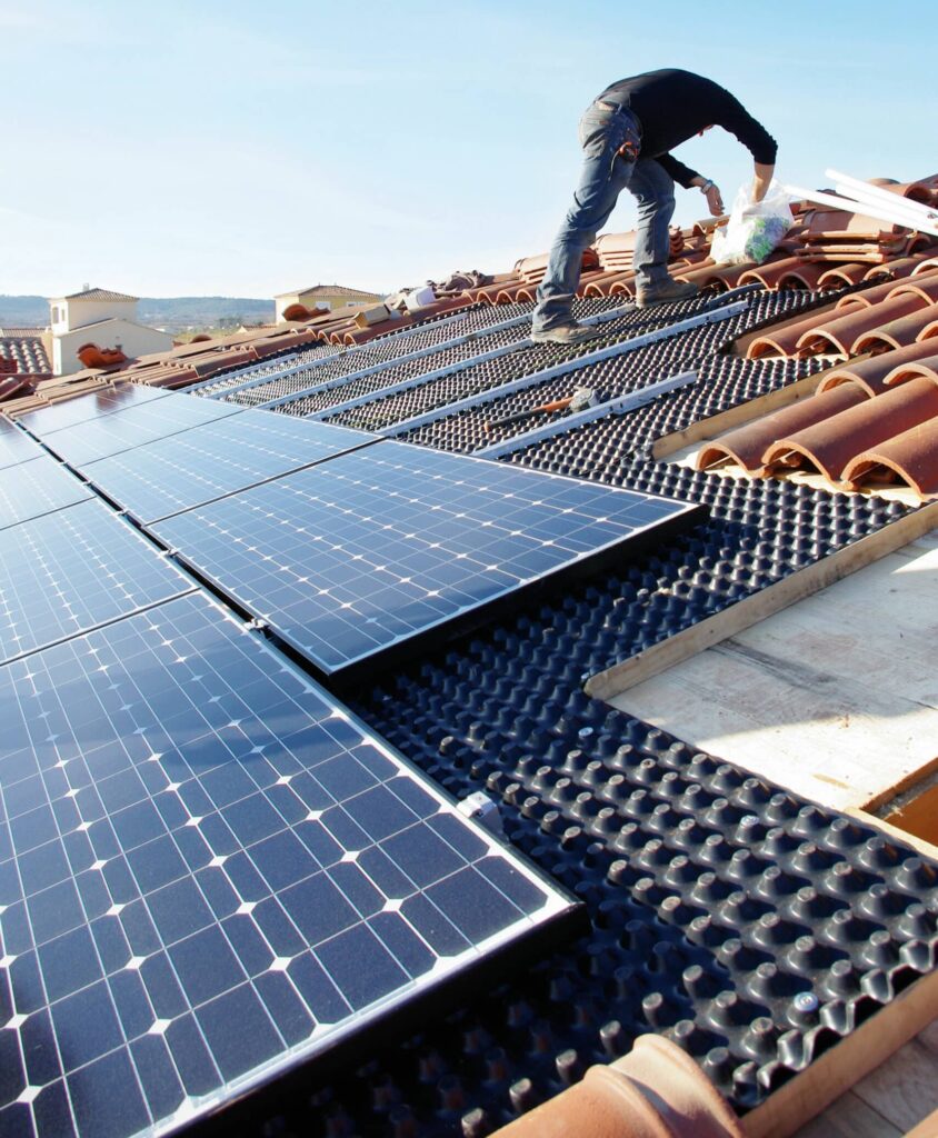 Panneaux solaires : installation écologique sur toit de tuiles Installateur posant des panneaux solaires photovoltaïques sur un toit en tuiles, avec une sous-couche noire texturée. Ciel bleu.