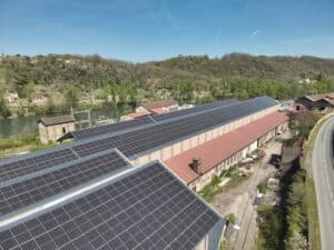 Vue aérienne d'installations industrielles au bord d'une rivière, avec de nombreux panneaux solaires sur les toits et un paysage boisé en arrière-plan.