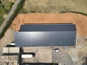 Vue aérienne d'un grand entrepôt industriel avec un toit en tôle ondulée gris foncé et argenté, près d'une route et d'une zone terreuse.