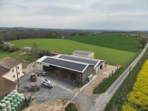 Ferme rurale avec toits photovoltaïques, ballots de paille filmés, tracteur et camion-grue sur cour, paysages vallonnés et champ de colza jaune.