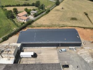 Vue aérienne d'une nouvelle grande toiture industrielle gris foncé avec ouvriers travaillant sur la sécurité du toit ; champ de foin et maisons rurales en arrière-plan.