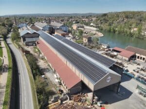 Vue aérienne d'un site industriel avec une grande toiture couverte de panneaux solaires et le logo "4 TOUT FER". Rivière et ville en arrière-plan.