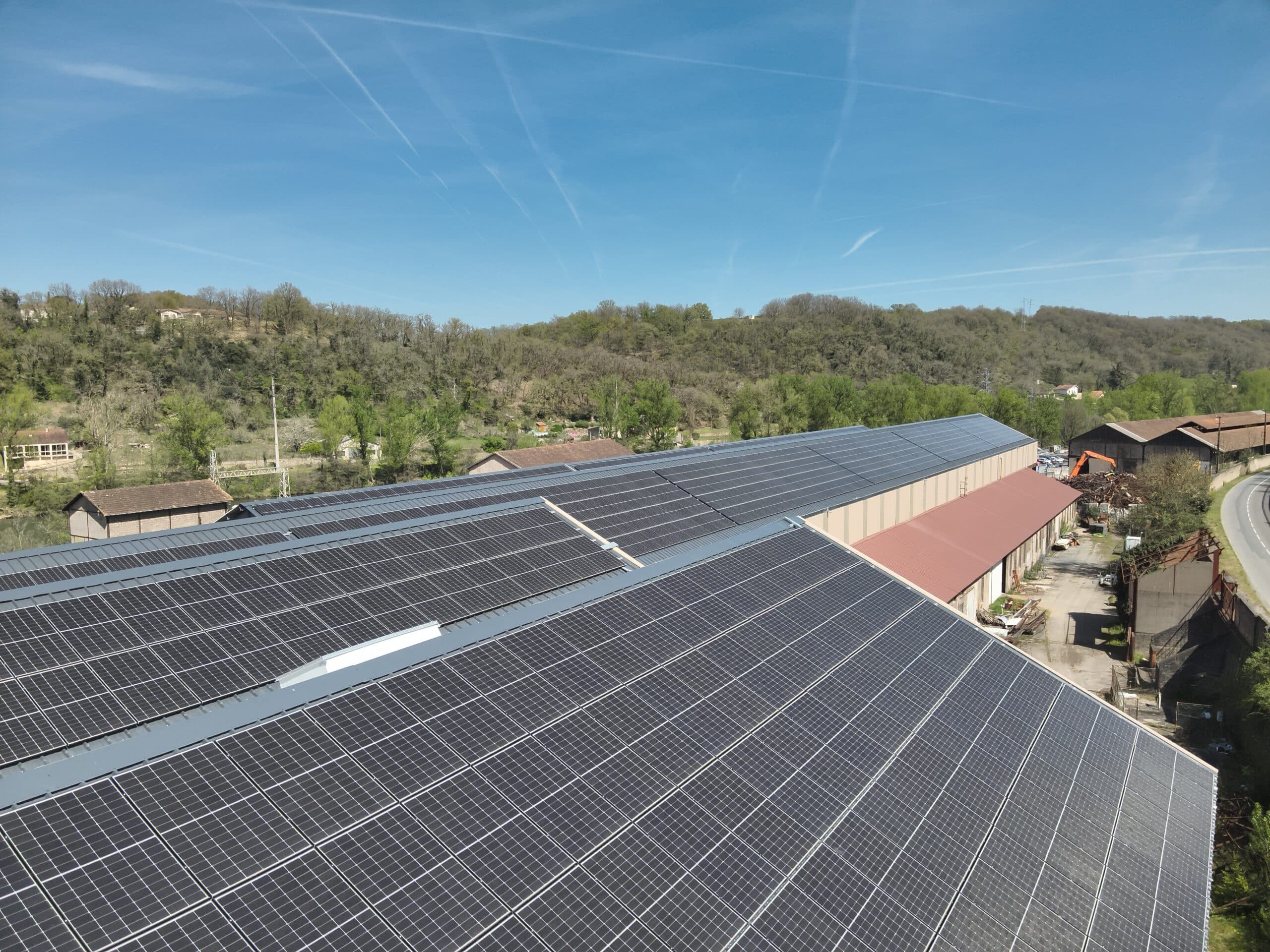 Toit industriel couvert de panneaux solaires, dominant un paysage vallonné et boisé par un ciel bleu clair avec traînées de condensation.