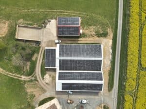 Vue aérienne de fermes avec multiples panneaux solaires sur toits rouges et blancs, bordées par une route et un champ de colza jaune.