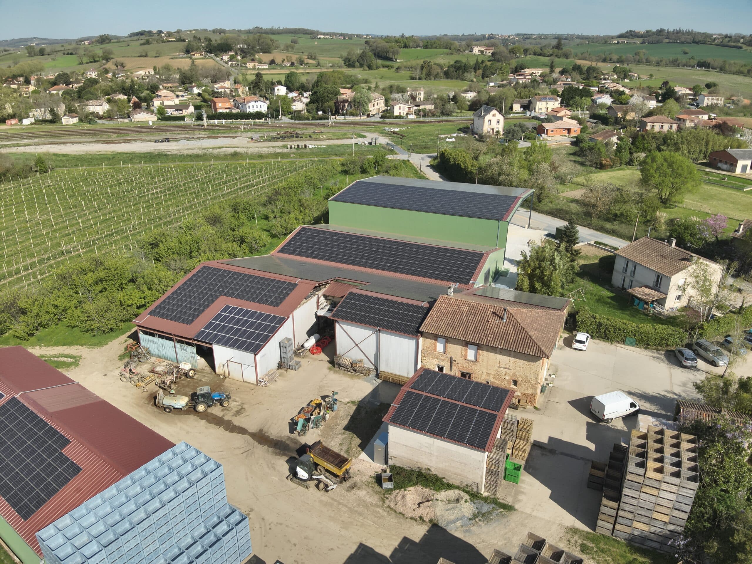 Vue aérienne d'une exploitation agricole avec de nombreux panneaux solaires sur les toits, des vignes, et un village vallonné en arrière-plan.