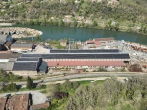Vue aérienne d'une usine avec toits recouverts de panneaux solaires, près d'une rivière et d'un barrage rocheux, avec habitations en zone boisée.